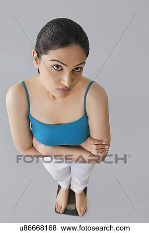 Portrait of worried young woman standing with arms crossed on weighing scale against gray background View Large Photo Image Stock Photo - Portrait of worried young woman standing with arms crossed on weighing scale against gray background . Fotosearch