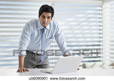 Portrait of young businessman standing by desk with laptop View Large Photo Image Stock Image - Portrait of young businessman standing by desk with laptop . Fotosearch