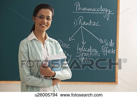 Picture - Portrait of young female teacher holding books against green board . Fotosearch