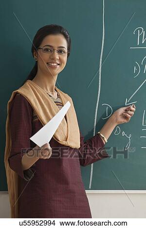 Picture - Portrait of young female teacher explaining a lesson while standing against chalkboard . Fotosearch