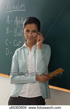 Picture - Portrait of young female teacher wearing glasses while leaning on chalkboard . Fotosearch