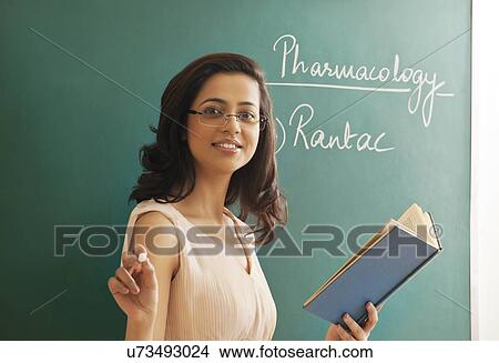 Picture - Portrait of young female teacher holding chalk and book against green board . Fotosearch