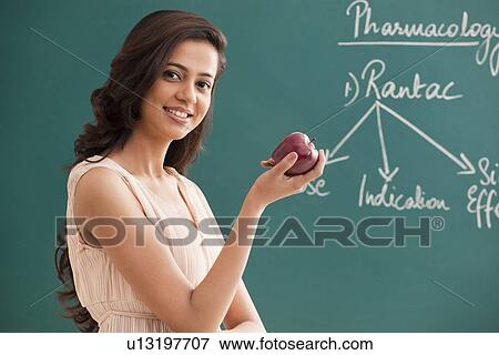 Stock Photo - Portrait of young female teacher holding an apple against green board . Fotosearch