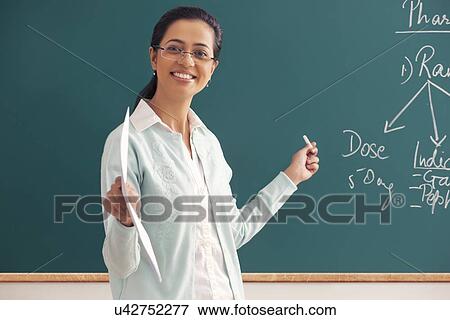 Portrait of young female teacher holding paper and chalk against green board View Large Photo Image Stock Photo - Portrait of young female teacher holding paper and chalk against green board . Fotosearch