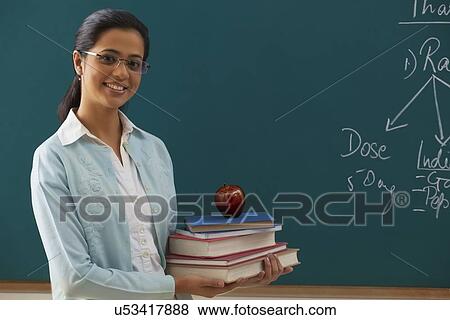 Stock Photo - Portrait of young female teacher holding stack of books against chalkboard . Fotosearch