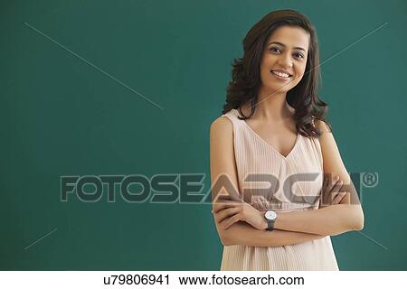 Stock Image - Portrait of young female teacher with arms crossed standing against green board . Fotosearch