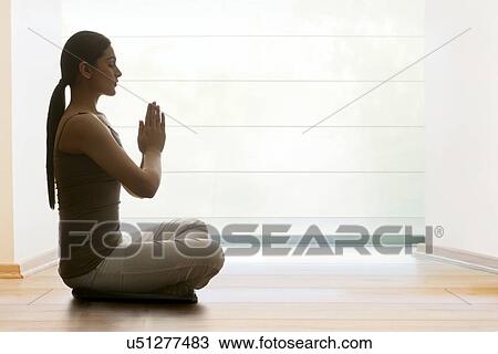 Stock Image - Profile shot of young woman with hands clasped meditating on hardwood floor. Fotosearch