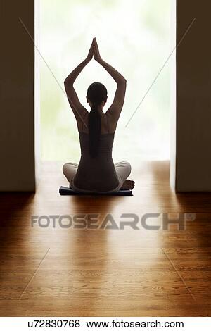 Stock Photo - Rear view of young woman meditating on hardwood floor while raising hands. Fotosearch