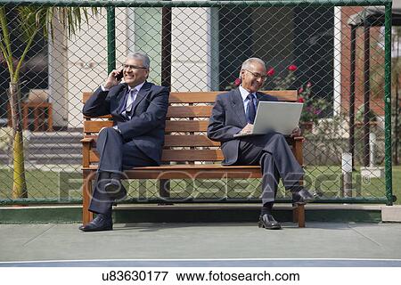 Senior businessman on phone call with other executive using laptop while sitting on bench in a tennis court View Large Photo Image Stock Photo - Senior businessman on phone call with other executive using laptop while sitting on bench in a tennis court . Fotosearch