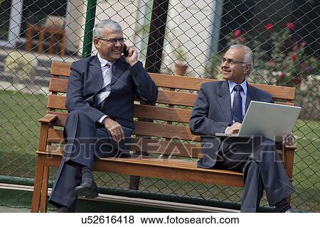 Stock Photo - Senior businessmen on bench looking at each other while using cell phone and laptop . Fotosearch