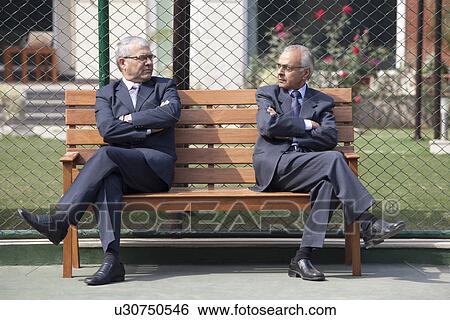 Stock Photograph - Senior male business executives staring at each other while sitting arms crossed on bench in tennis court . Fotosearch