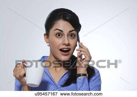 Stock Photo - Shocked businesswoman with coffee mug answering call against gray background. Fotosearch