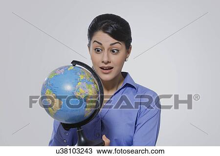 Stock Image - Shocked young businesswoman looking at globe against gray background. Fotosearch