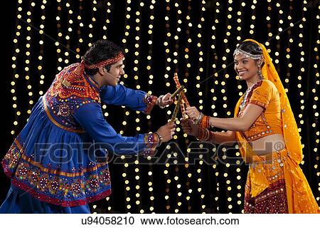 Stock Image - Side view of young couple in traditional wear performing Dandiya Raas against neon lights. Fotosearch