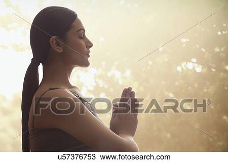 Stock Image - Side view of young woman with hands clasped meditating against trees. Fotosearch