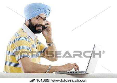 Stock Photography - Sikh man talking on the phone while working on a laptop . Fotosearch