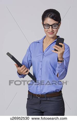 Stock Image - Smiling businesswoman using mobile phone while holding laptop against gray background. Fotosearch