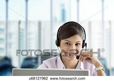 Stock Image - Smiling businesswoman wearing headset and using laptop in office . Fotosearch