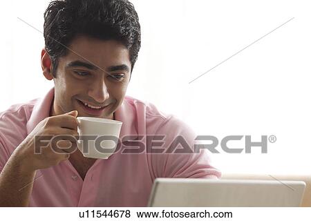 Smiling man having coffee while using laptop View Large Photo Image Stock Photo - Smiling man having coffee while using laptop . Fotosearch