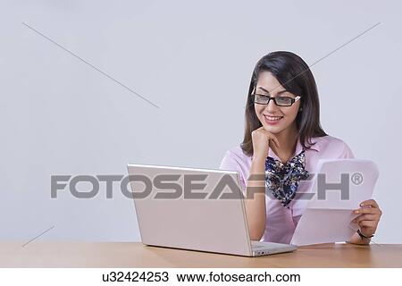 Stock Image - Smiling young businesswoman working on laptop at desk in office. Fotosearch