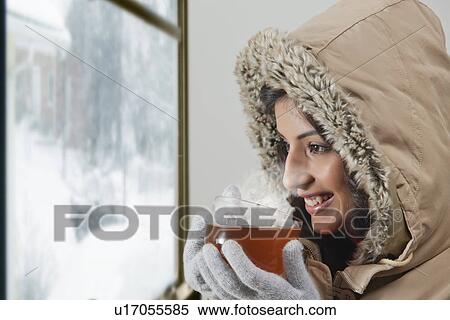 Stock Photography - Smiling young woman looking through window and drinking tea . Fotosearch