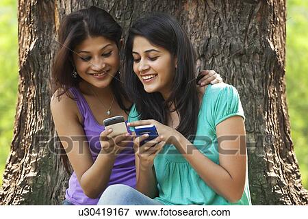 Stock Photo - Smiling young women sitting against tree trunk using cell phone . Fotosearch