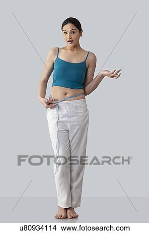 Picture - Surprised young woman measuring waist isolated over gray background . Fotosearch