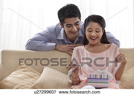 Stock Photo - Surprised young woman unwrapping gift with man at home . Fotosearch