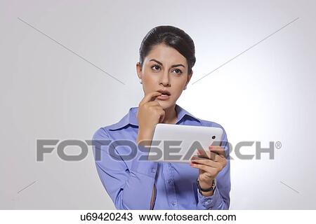 Stock Image - Thoughtful businesswoman with finger in mouth holding digital tablet over gray background. Fotosearch