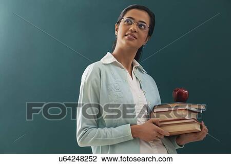 Stock Image - Thoughtful female teacher carrying stack of books and an apple against chalkboard . Fotosearch