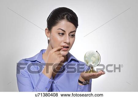 Stock Photo - Thoughtful Indian businesswoman exploring globe against gray background. Fotosearch