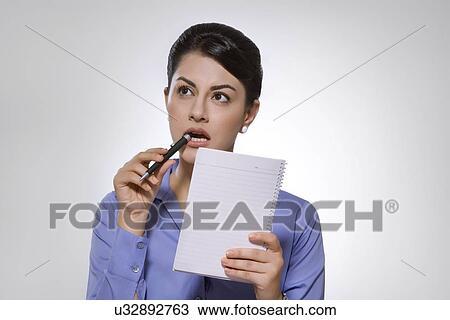 Stock Image - Thoughtful Indian businesswoman with note pad and pen against gray background. Fotosearch