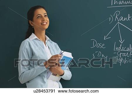 Stock Image - Thoughtful young female teacher holding books and glasses against green board . Fotosearch