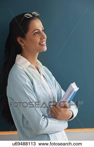 Thoughtful young female teacher holding books against green board View Large Photo Image Stock Image - Thoughtful young female teacher holding books against green board . Fotosearch