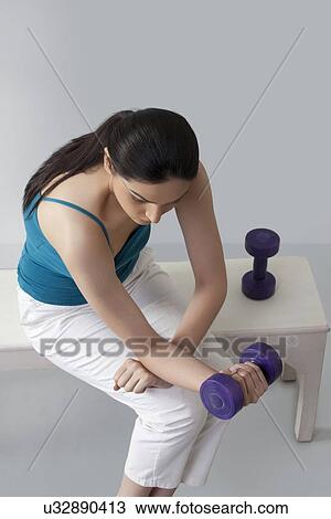 Stock Image - Woman lifting dumbbells while sitting on bench against gray background . Fotosearch