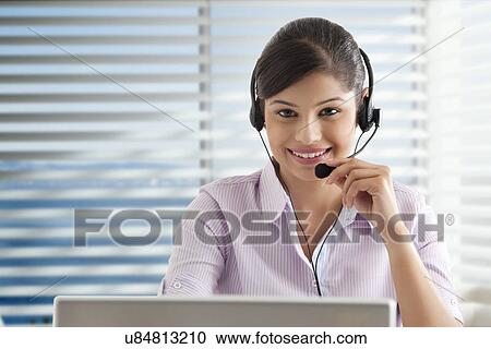 Stock Image - Young businesswoman using laptop in office, wearing headset . Fotosearch