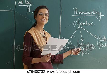 Stock Image - Young female teacher explaining a lesson while standing against chalkboard . Fotosearch