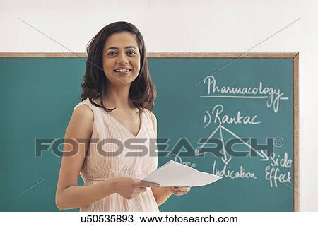 Stock Image - Young female teacher holding papers against green board . Fotosearch