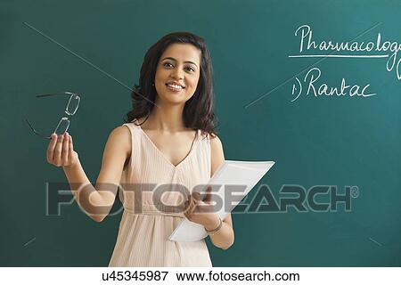Stock Photo - Young female teacher looking away while holding glasses and file against green board . Fotosearch