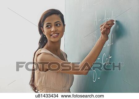 Young female teacher writing on green board while looking away View Large Photo Image Picture - Young female teacher writing on green board while looking away . Fotosearch