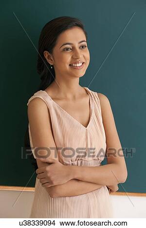 Picture - Young happy female teacher with arms crossed standing against green board . Fotosearch