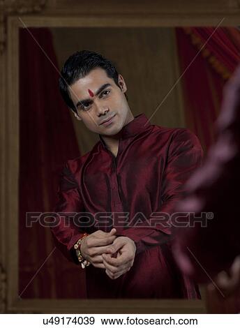 Stock Photo - Young Indian bridegroom looking at self in mirror while getting dressed . Fotosearch