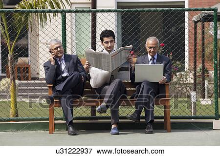 Stock Photo - Young male executive reading newspaper while sitting among senior executives on bench . Fotosearch