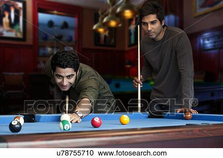 Stock Image - Young man aiming at cue ball while friend standing by pool table . Fotosearch