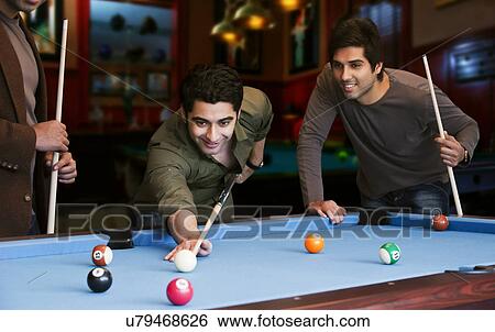 Stock Photograph - Young man aiming at cue ball while friends standing by pool table . Fotosearch