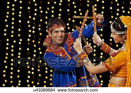 Picture - Young man performing Dandiya Rass with woman against neon lights. Fotosearch