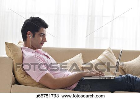 Stock Image - Young man using laptop while listening to music on sofa . Fotosearch