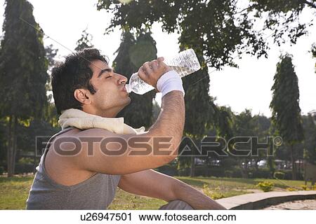 Stock Image - Young man with towel round shoulders drinking water after workout . Fotosearch