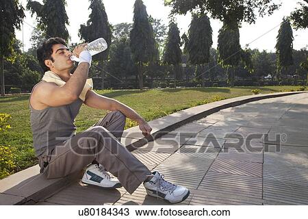 Stock Image - Young man with towel round shoulders drinking water after workout . Fotosearch