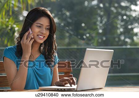 Stock Image - Young woman answering phone call while working on laptop . Fotosearch